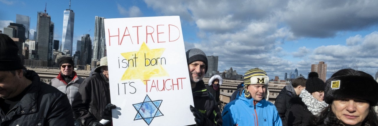 Photo of people marching on a bridge under a blue sky, man holding a poster sign that says 'hatred isn't born it's taught' with a Jewish star on the poster