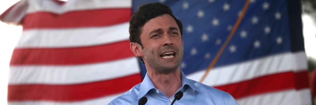 Senator Jon Ossoff (D-GA) in a blue shirt speaking into a microphone in front of a US Flag