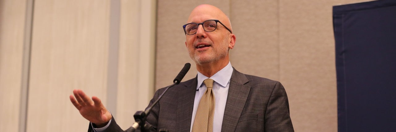 Image of AJC CEO Ted Deutch in suit and tie, speaking, wearing glasses, in front of a clear lectern