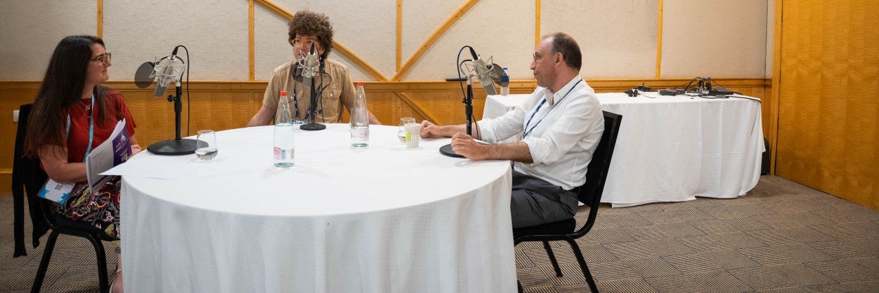 Three people sitting around a round table with a white tablecloth, one woman and two men, sitting in front of microphones