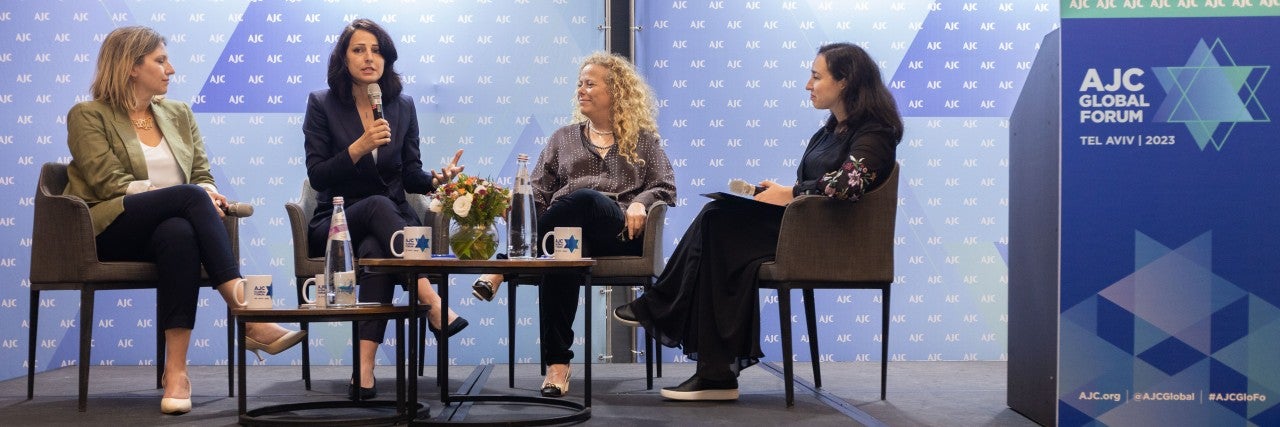 four people, Aviva Steinberger, Gadeer Kamal-Mreeh, Ayelet Nahmias-Verbin, Reva Gorelick, sitting on a black stage in front of a blue step and repeat background with the American Jewish Committee (AJC) logo, two tables with water bottles on top, and on the right side of the stage is a blue AJC-branded podium with a microphone