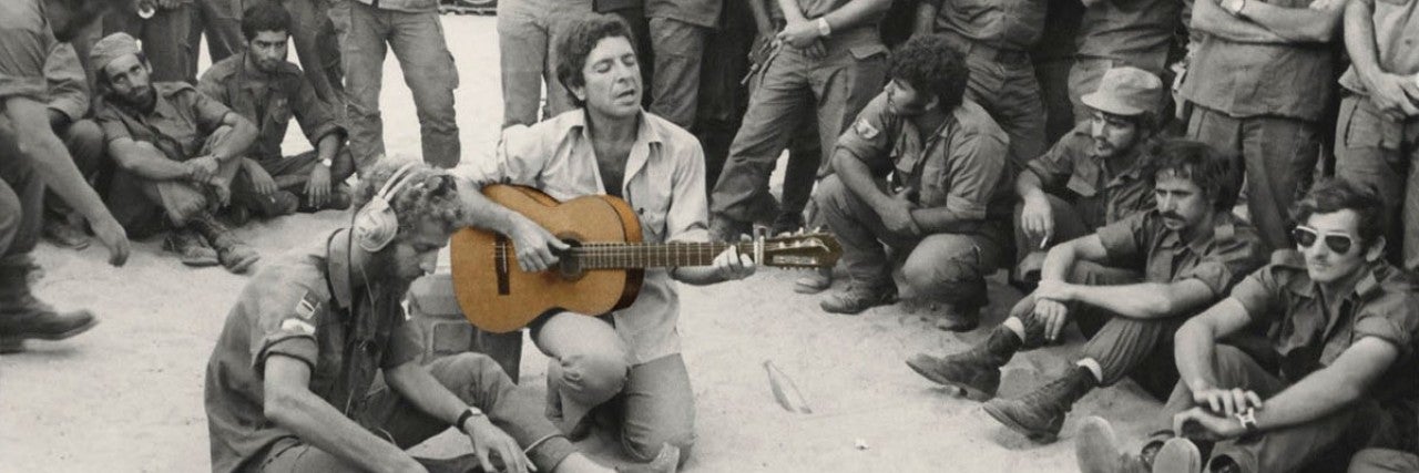 Leonard Cohen in the Sinai Desert in October 1973, on a concert tour. Sitting on the ground, holding a guitar. 