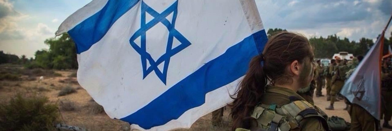 Image of large Israeli flag flying over a blue sky, a soldier Mai Gutman holding it