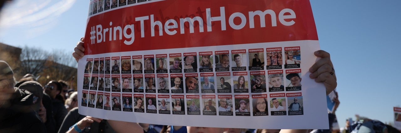 someone holding up a large sign that covers most of the frame. the sign is a mosaic of images of over 200 people kidnapped by hamas. there is a red banner going through the middle that reads in white text, #BringThemHome. the background is a blue sky.