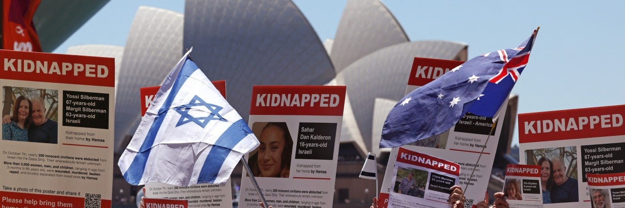 crowd of people at a rally for israel, holding signs of people taken hostage by hamas, in front of the opera house in sydney, australia. austrailian and israeli flags are being held up, flowing in the wind.