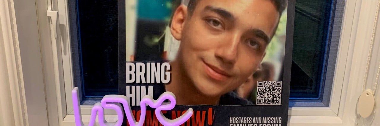 Edan Alexander hostage missing persons poster. red text reading Edan Alexander in Hebrew and English, 19 crossed out and 20 is drawn on. Below is a photo of a smiling young man. The poster is leaning on a windowsill and a small purple "love" sculpture is placed in front of the poster.