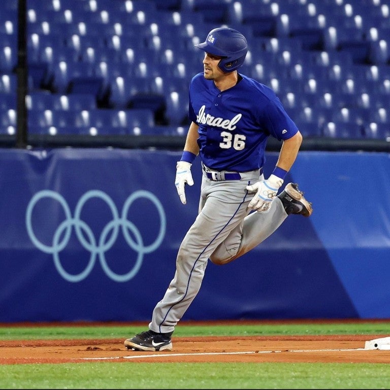 Photo of baseball player ryan lavarnway with a jersery that reads "Israel 36" with olympic rings in the background, on a baseball field, running