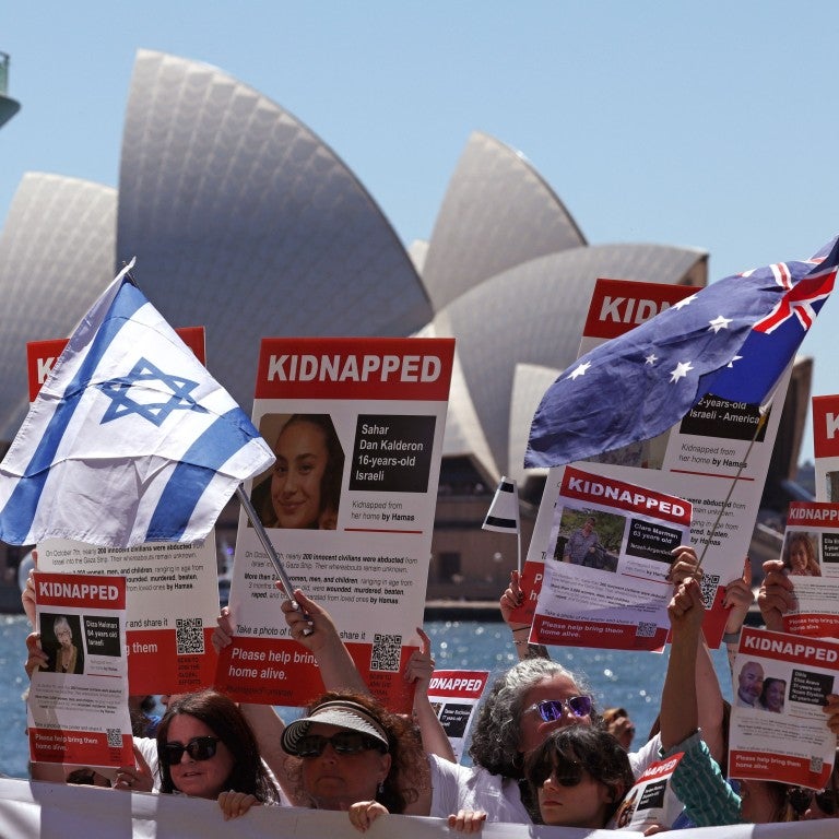 crowd of people at a rally for israel, holding signs of people taken hostage by hamas, in front of the opera house in sydney, australia. austrailian and israeli flags are being held up, flowing in the wind.